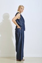 A model stands in profile in a modern studio wearing a sophisticated denim outfit. The light