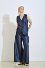 A fashionable woman stands confidently in a sleek denim vest and wide-leg pants. Her accessories