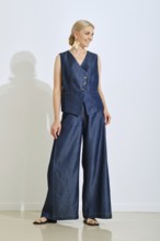 A model is wearing a chic denim vest and wide-legged pants in a well-lit studio. She poses