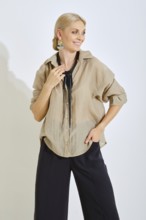 A woman is standing with a smile, dressed in a light beige shirt and dark pants. She wears a