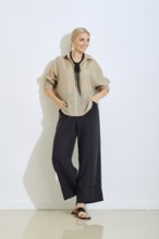 A woman stands confidently in a relaxed beige shirt and black pants, hands in pockets, on a clean