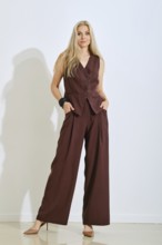A model stands confidently in a brown vest and matching pants. Her long hair flows down as she