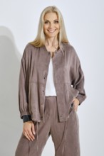 A smiling woman with long blonde hair is posing confidently in a soft brown casual outfit. She is