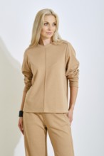 A confident woman stands elegantly in a bright studio, wearing a simple beige top and matching