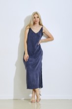 A woman stands gracefully against a plain white wall, wearing a long, sleeveless dress in a deep