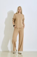 A cheerful woman is posing indoors wearing a light beige two-piece outfit that includes a