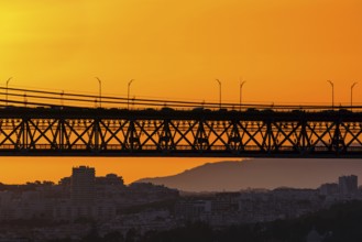 Bridge spans city skyline, silhouetted against vivid orange sunset. Soft city outlines and distant