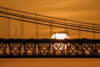 Glowing sun setting behind 25 de Abril bridge, Lisbon, Portugal, suspension cables, cars and bridge
