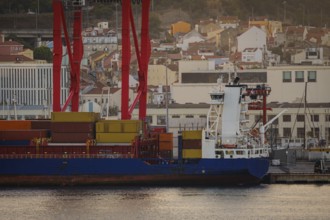 Cargo ship loaded with multicolored containers docked at Lisbon port, red cranes and city vibrant
