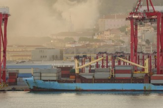 Blue container ship docked at Lisbon port, red cranes and stacks of shipping containers on deck,