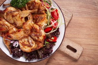 Baked quail, served on a plate with crispy lettuce leaves, fresh tomatoes and herbs, on a wooden