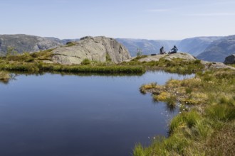 Two people sitting on a rock by a mountain lake, above the Preikestolen hiking trail and high above