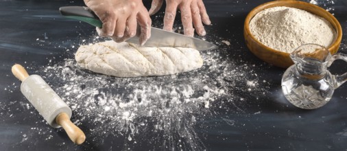 Hands of a baker scoring raw bread dough with a knife on a flour-dusted surface, preparing fresh