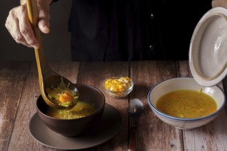 Woman serving chicken soup with a wooden spoon in a brown bowl on a wooden table