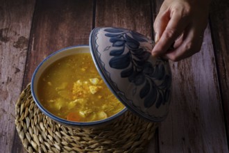 Woman opening the lid of a tureen with chicken noodle soup and vegetables on a wooden table