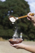 Hands sifting white flour from a wooden spoon into a rustic ceramic bowl, preparing ingredients for