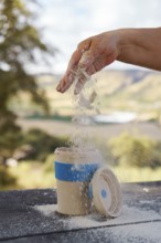 Hand pouring white powder from hand into a reusable portable mug on a wooden table, enjoying
