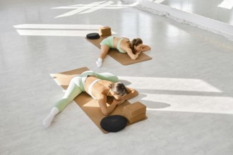 Two women are engaged in a stretching session, using mats and blocks in a well-lit studio space.