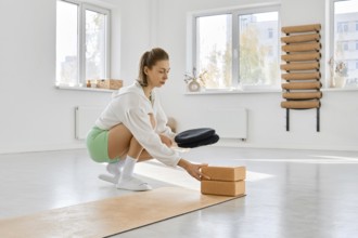 A woman squats on a yoga mat in a spacious studio. She is using stretching blocks to enhance her