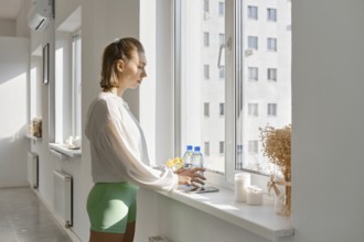 A young woman stands by a large window, feeling the warm sunlight in a bright indoor space. She