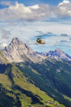 A helicopter flies over a mountainous region under a cloudy sky