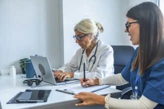 Two medical professionals, a senior doctor typing on a laptop and a younger nurse taking notes on a