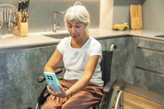 Senior woman in a wheelchair video calling on a smartphone from a modern kitchen, engaging in