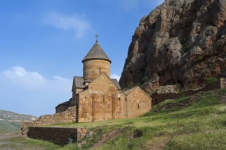 A stone monastery building rising on rough rock walls, surrounded by blue skies and green meadows,