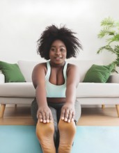 A woman is doing yoga on a mat in front of a couch. She is wearing a tank top and gray pants, AI