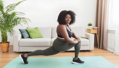 A woman is doing a yoga pose on a mat in a living room. She is wearing a green top and green pants,