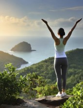 A woman is standing on a mountain top, looking out over the ocean. She is wearing a white tank top