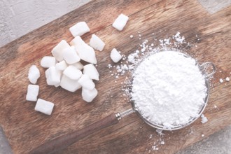 A strainer with powdered sugar, on a wooden cutting board, with pieces of white sugar