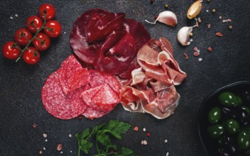 Assorted dried meat, fresh tomatoes and olives spread out on a dark surface, food composition, top