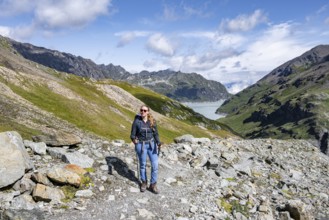 Person posing on rocky path with background of mountains and a lake under blue sky, Lac des Dix