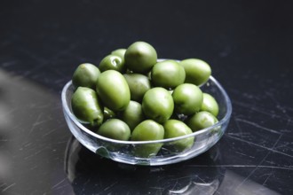 A clear bowl holds a generous pile of green olives. The olives are glossy and vibrant, resting on a