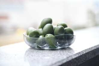 A bowl filled with green olives on a smooth granite surface. The sunlight reflects off the glass