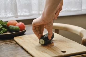 A hand holds a cucumber while cutting it on a wooden board. Fresh tomatoes and greens are arranged