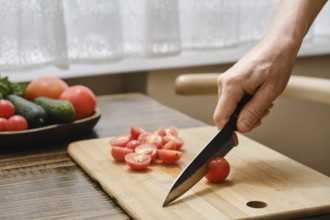 A hand holds a knife, skillfully slicing fresh cherry tomatoes on a wooden cutting board. Various