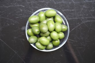 Overhead view of a bowl filled with green olives on a black surface. The olives are vibrant and