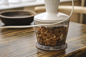 A food processor is filled with walnuts on a wooden table. Natural light filters through a nearby