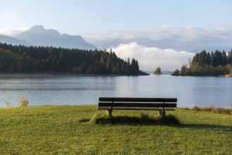 A lake with a bench in the foreground, surrounded by forests and mountains under a blue sky,
