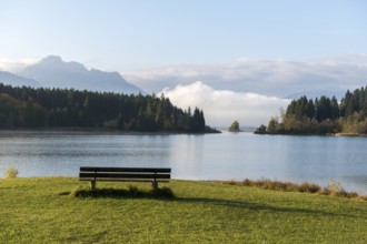 A bench stands in front of a quiet lake, surrounded by mountains and forests in clear air,