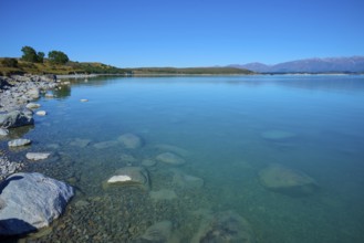 Shore with stones in clear water surrounded by vast countryside and mountains, Lake Pukaki