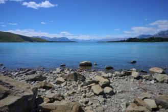 Clear water gently washes against rocky shoreline surrounded by high mountains under bright blue
