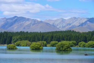A lake with small green islands and wooded slopes against a mountain range and blue sky, Lake