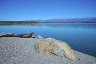 Rocky shore on a calm blue lake with views of distant mountains, Lake Pukaki Viewpoint, Pukaki,