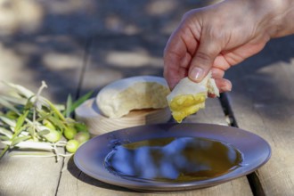 Hand dipping fresh bread into a plate of extra virgin olive oil on a rustic wooden table, with