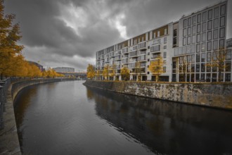 Modern apartment block on the river with yellow autumn trees, dramatic sky reflected in calm water,