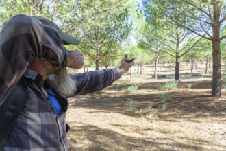 Man with beard and plaid shirt aiming a slingshot, focusing intently on a target in a pine forest