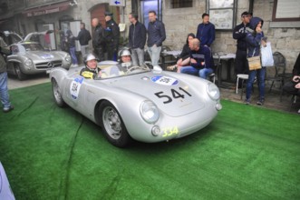 Silver Porsche 550 Spyder with two helmeted drivers drives across the green carpet at the rally,